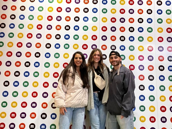 Mayra Rivera, MS, Buchan Lab, Vanessa and Lucas pose in front of M&M themed backboard at the conference