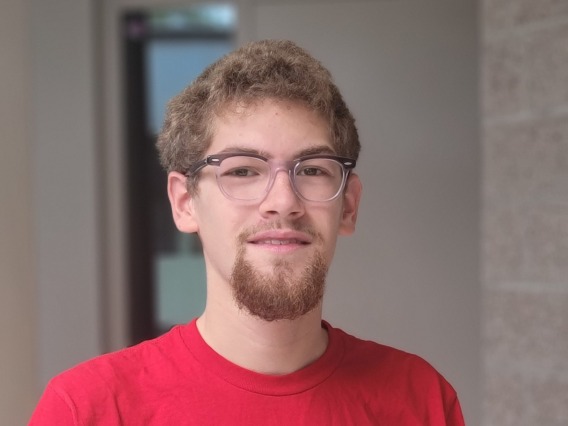 Ben Augustine, wearing a red t-shirt and glasses, smiling towards the camera