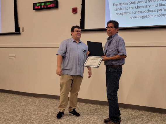 Hollis Whitewater (right) receiving the Dr. Harold McNair Staff Award from Craig Aspinwall (left). Both standing in front of a projector screen and a wall clock on the top left.