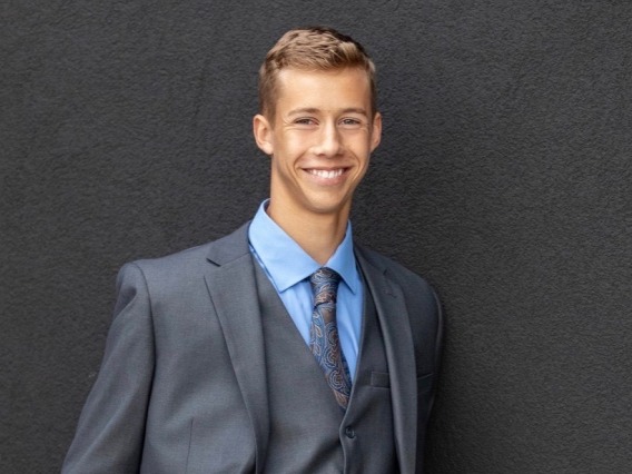 Evan Thomas wearing a dark suit, smiling towards the camera in front of a black back drop
