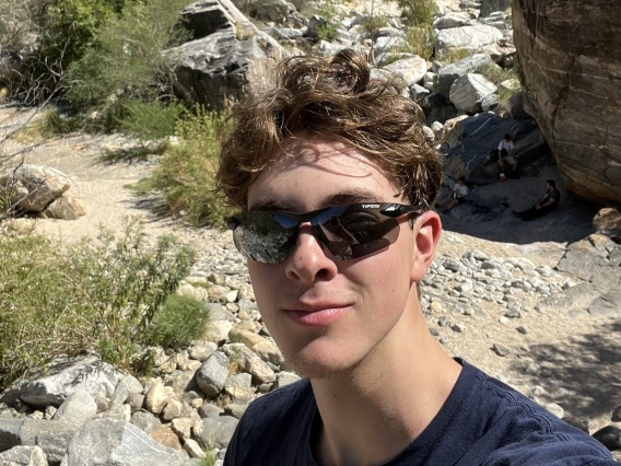 Jackson Taylor in the desert wearing black shades and a navy blue tee smiling towards the camera