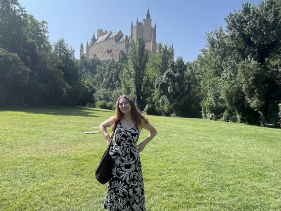 Olivia Seagraves in Spain, wearing a black and white dress standing on an open grass field, there is a castle and trees in the background.