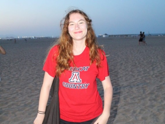 Olivia Seagraves at the beach, smiling towards the camera wearing a red University of Arizona shirt and a black tote bag.