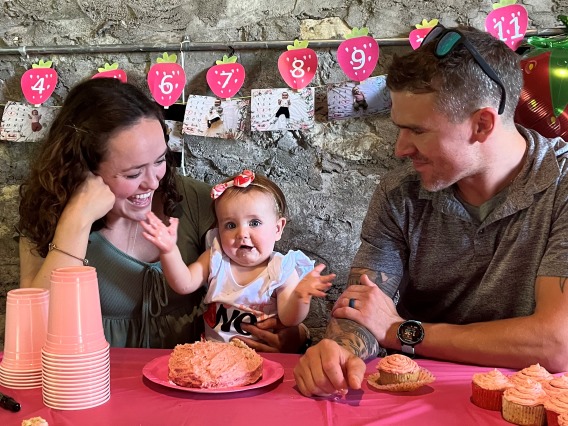 Kirsten Caldwell with baby and partner sitting at a table in a restaurant. Baby with open arms facing the camera
