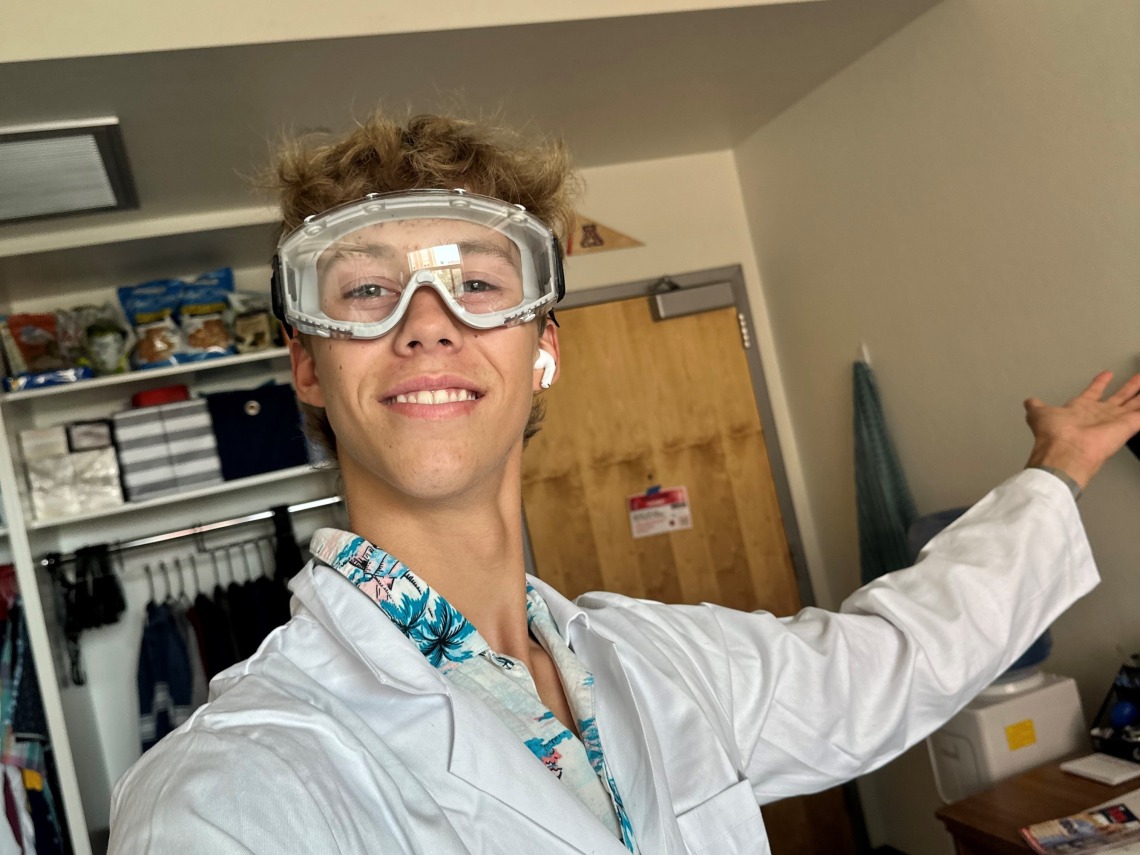 Evan Thomas wearing a lab coat and safety goggles with one arm extended to the right, smiling towards the camera