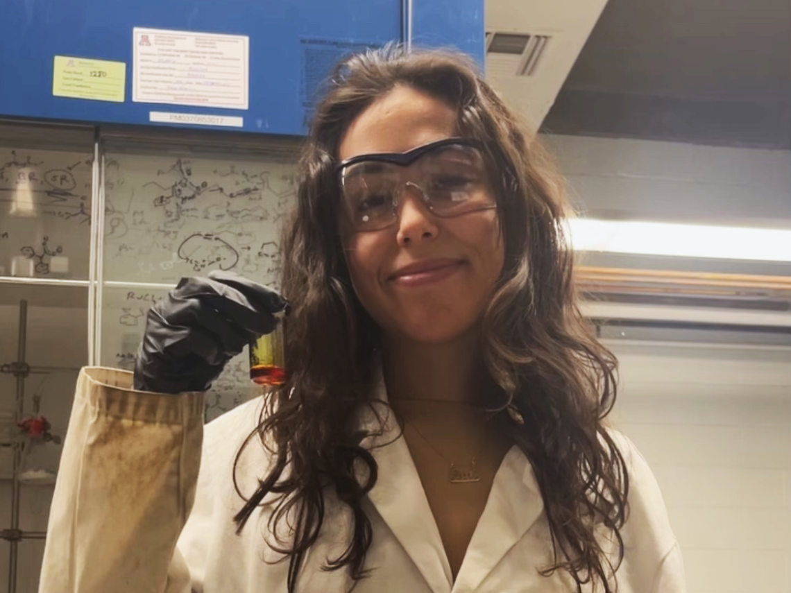 Gal Melman smiling towards the camera and standing in a lab. Wearing a lab coat, black gloves, and safety goggles, holding a beaker with a red substance inside.
