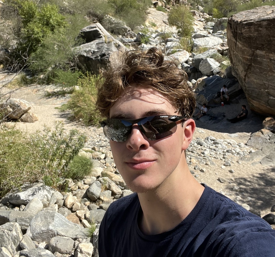 Jackson Taylor in the desert wearing black shades and a navy blue tee smiling towards the camera